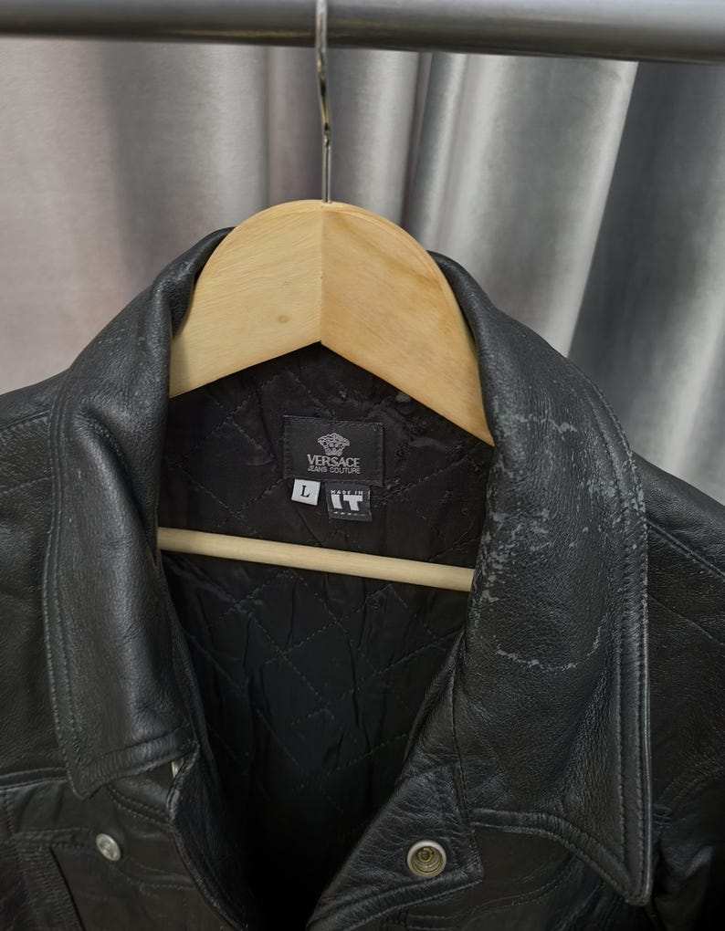 Puede incluir: Una chaqueta de cuero negro con forro interior acolchado y cuello en punta. La etiqueta interior muestra el logotipo de Versace Jeans Couture, una etiqueta de talla L y una etiqueta Made in IT. El cuero muestra signos visibles de desgaste.