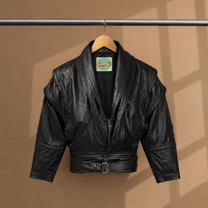 May include: A black leather jacket featuring a wide shawl collar, padded shoulders, and a belted waist with a silver-toned buckle. The jacket has a central zipper closure and a circular label at the inner neckline with a globe graphic.