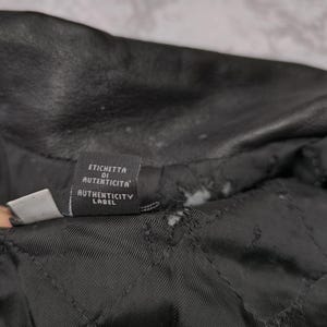 Puede incluir: El interior de una prenda de cuero negra con forro acolchado y una peque&ntilde;a etiqueta de tela rectangular. La etiqueta muestra el texto ETICHETTA DI AUTENTICITA y AUTHENTICITY LABEL en letras blancas sobre un fondo negro.