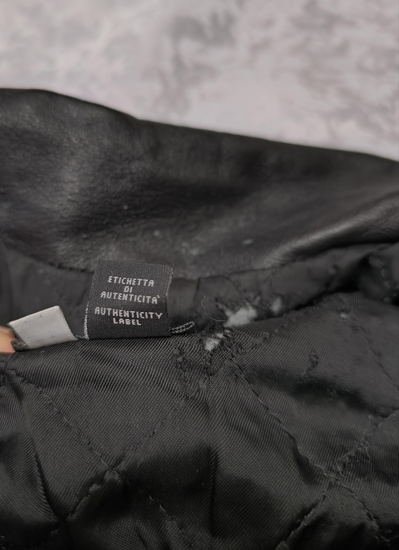 Puede incluir: El interior de una prenda de cuero negra con forro acolchado y una peque&ntilde;a etiqueta de tela rectangular. La etiqueta muestra el texto ETICHETTA DI AUTENTICITA y AUTHENTICITY LABEL en letras blancas sobre un fondo negro.