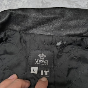Puede incluir: Una chaqueta negra Versace Jeans Couture con forro interior acolchado. Una etiqueta de tela negra est&aacute; cosida en el interior, mostrando el logotipo de la marca, una etiqueta de talla L y una etiqueta que dice Made in IT.