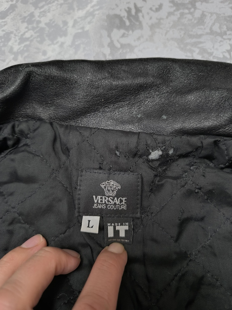 Puede incluir: Una chaqueta negra Versace Jeans Couture con forro interior acolchado. Una etiqueta de tela negra est&aacute; cosida en el interior, mostrando el logotipo de la marca, una etiqueta de talla L y una etiqueta que dice Made in IT.