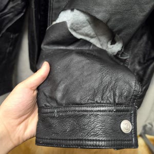 Puede incluir: Una manga de chaqueta de cuero negro con superficie texturizada, un pu&ntilde;o abotonado con un broche de metal plateado y un forro interior gris visible a trav&eacute;s de un desgarro en el material de cuero cerca de la zona del brazo superior.