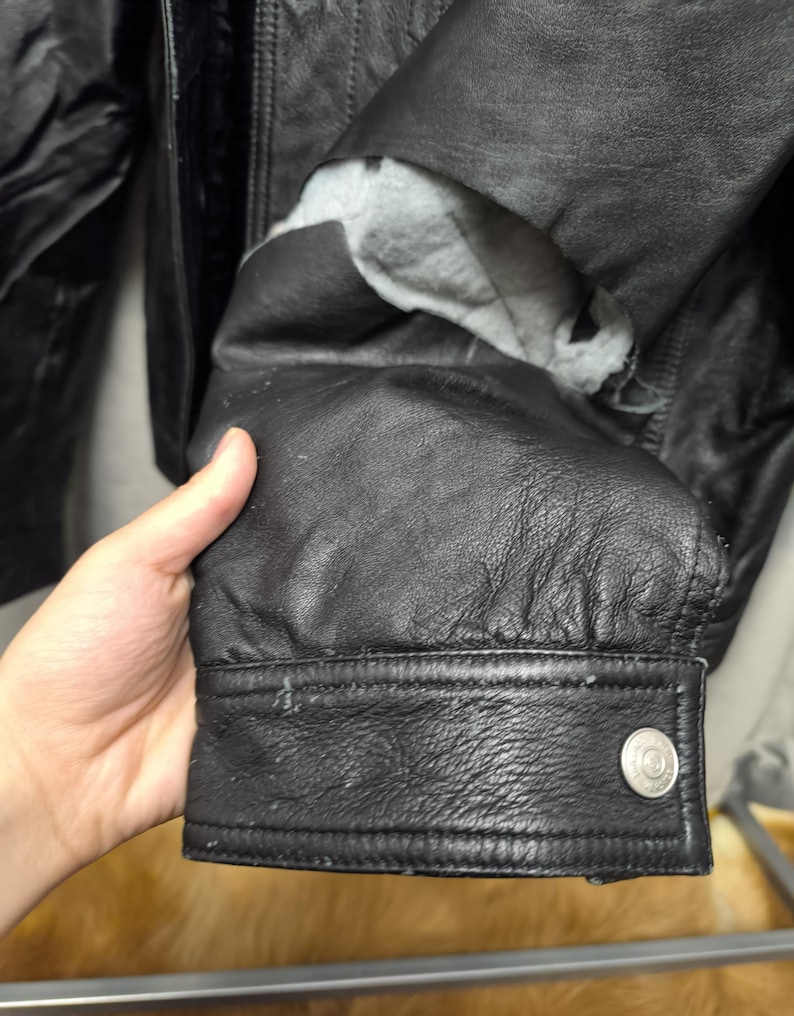 Puede incluir: Una manga de chaqueta de cuero negro con superficie texturizada, un pu&ntilde;o abotonado con un broche de metal plateado y un forro interior gris visible a trav&eacute;s de un desgarro en el material de cuero cerca de la zona del brazo superior.