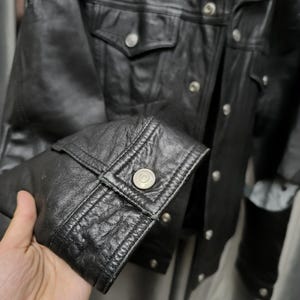 Puede incluir: Una chaqueta de cuero negro con acabado texturizado, cuello en punta y mangas largas con pu&ntilde;os abotonados. La parte delantera de la chaqueta incluye un cierre de botones plateados met&aacute;licos y dos bolsillos en el pecho con solapas abotonadas.