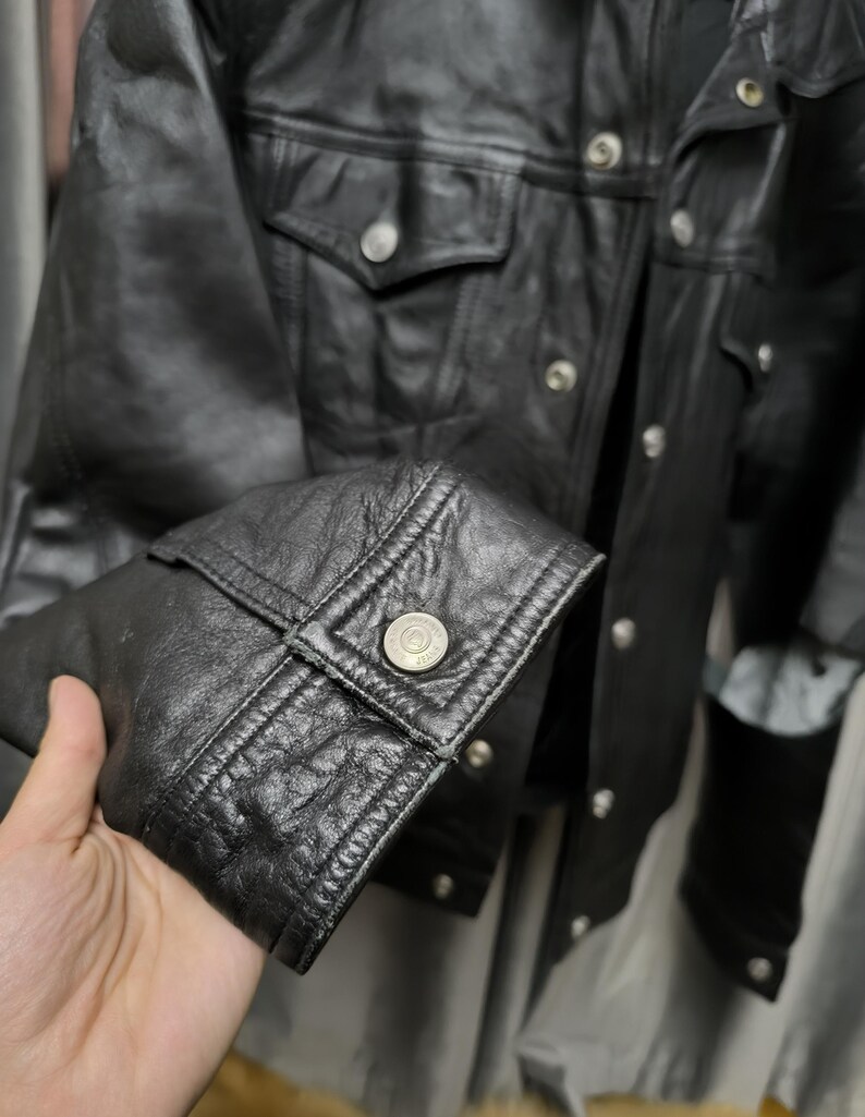 Puede incluir: Una chaqueta de cuero negro con acabado texturizado, cuello en punta y mangas largas con pu&ntilde;os abotonados. La parte delantera de la chaqueta incluye un cierre de botones plateados met&aacute;licos y dos bolsillos en el pecho con solapas abotonadas.