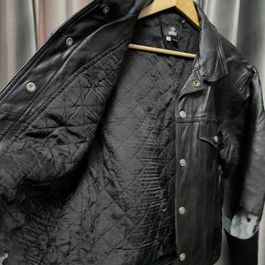 Puede incluir: Una chaqueta de cuero negro con forro interior acolchado en negro, cuello cl&aacute;sico y botones de presi&oacute;n de metal plateado en la parte delantera. La chaqueta se muestra en una percha de madera e incluye una etiqueta de marca visible en el cuello.