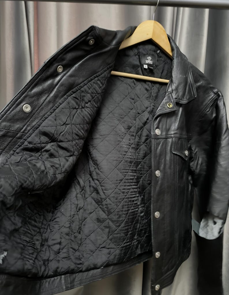 Puede incluir: Una chaqueta de cuero negro con forro interior acolchado en negro, cuello cl&aacute;sico y botones de presi&oacute;n de metal plateado en la parte delantera. La chaqueta se muestra en una percha de madera e incluye una etiqueta de marca visible en el cuello.