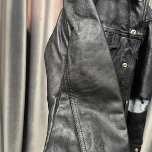 Puede incluir: Una chaqueta de cuero negro con cuello estructurado, mangas largas con costuras visibles y una tapeta frontal asegurada con botones de presi&oacute;n de metal plateado. La chaqueta tiene una textura suave y mate con un corte cl&aacute;sico.