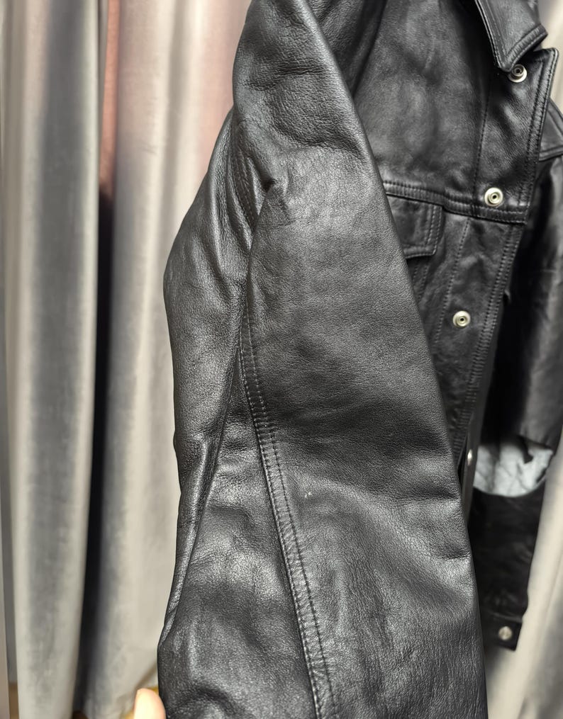 Puede incluir: Una chaqueta de cuero negro con cuello estructurado, mangas largas con costuras visibles y una tapeta frontal asegurada con botones de presi&oacute;n de metal plateado. La chaqueta tiene una textura suave y mate con un corte cl&aacute;sico.
