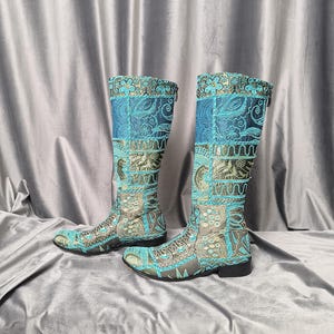 Op de afbeelding: Een paar kniehoge laarzen met een patchworkontwerp van blauwgroene en gouden stof. De laarzen zijn versierd met ingewikkelde bloemen- en wervelpatronen die met blauwgroene draad op een goudkleurige basis zijn geborduurd, met zwarte zolen en hakken.