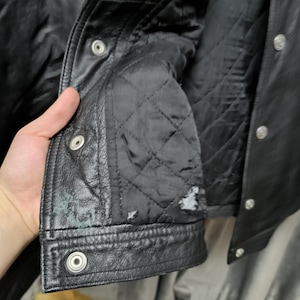 Puede incluir: Una chaqueta de cuero negro con forro interior acolchado, botones de presi&oacute;n de metal plateado a lo largo de la solapa frontal y desgaste visible con peque&ntilde;as &aacute;reas de p&eacute;rdida de material en la tela acolchada interior inferior.