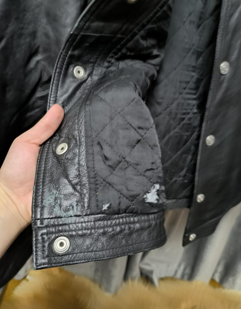 Puede incluir: Una chaqueta de cuero negro con forro interior acolchado, botones de presi&oacute;n de metal plateado a lo largo de la solapa frontal y desgaste visible con peque&ntilde;as &aacute;reas de p&eacute;rdida de material en la tela acolchada interior inferior.
