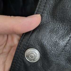 Puede incluir: Una chaqueta de cuero negro con un bot&oacute;n de metal plateado grabado con el texto Versace Jeans Couture y un logotipo central. La chaqueta presenta una superficie de grano texturizado y costuras visibles a lo largo del borde de la solapa frontal.