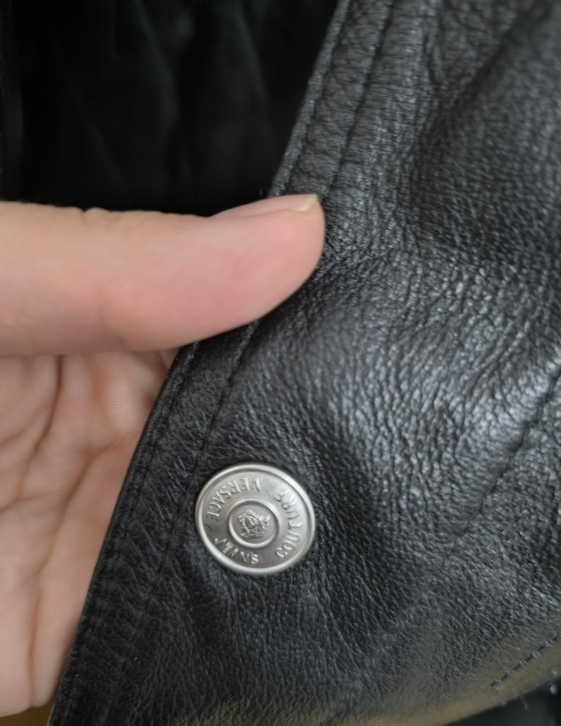 Puede incluir: Una chaqueta de cuero negro con un bot&oacute;n de metal plateado grabado con el texto Versace Jeans Couture y un logotipo central. La chaqueta presenta una superficie de grano texturizado y costuras visibles a lo largo del borde de la solapa frontal.