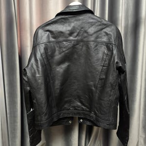Puede incluir: Una chaqueta de cuero negro vista desde atr&aacute;s, con cuello cl&aacute;sico, mangas largas y costuras visibles en los hombros y el panel posterior. La chaqueta est&aacute; confeccionada con un acabado oscuro y liso, e incluye un dobladillo con botones de presi&oacute;n.