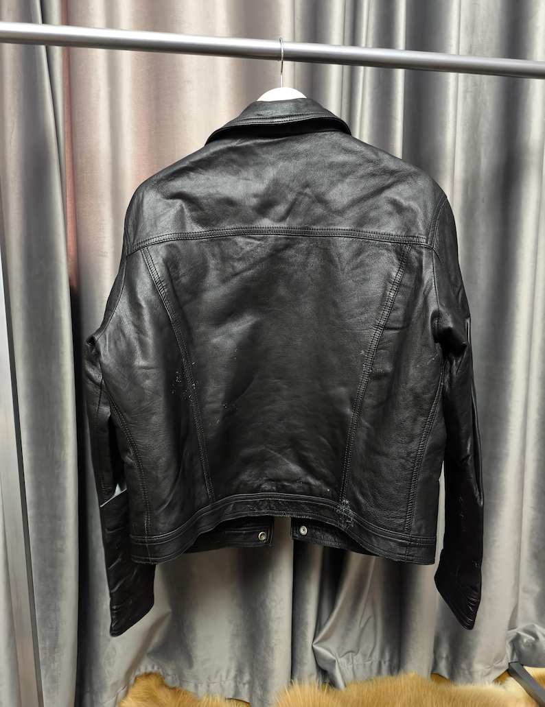 Puede incluir: Una chaqueta de cuero negro vista desde atr&aacute;s, con cuello cl&aacute;sico, mangas largas y costuras visibles en los hombros y el panel posterior. La chaqueta est&aacute; confeccionada con un acabado oscuro y liso, e incluye un dobladillo con botones de presi&oacute;n.