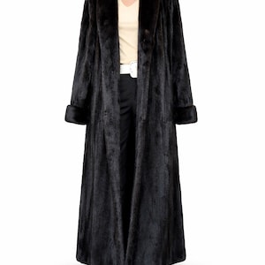 May include: A full-length black fur coat with a wide collar and cuffs. The coat is displayed on a mannequin, revealing a cream-colored top and black pants. A white belt cinches the waist.