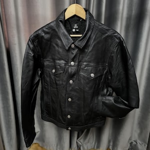 Puede incluir: Una chaqueta de cuero negro con cuello cl&aacute;sico, mangas largas y cierre frontal de botones con herrajes plateados. La prenda incluye dos bolsillos en el pecho con solapas abotonadas y una etiqueta de la marca Versace en el cuello interior.