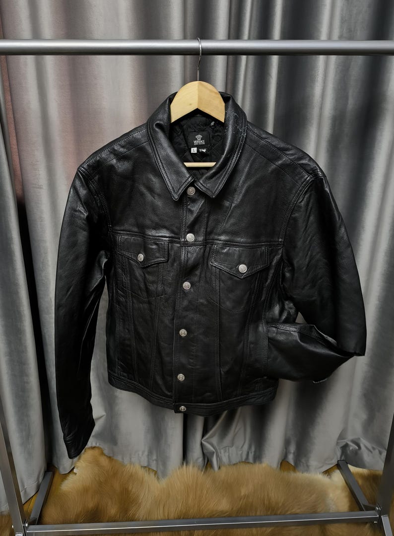 Puede incluir: Una chaqueta de cuero negro con cuello cl&aacute;sico, mangas largas y cierre frontal de botones con herrajes plateados. La prenda incluye dos bolsillos en el pecho con solapas abotonadas y una etiqueta de la marca Versace en el cuello interior.