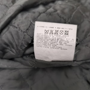 Puede incluir: Un interior de tela acolchada gris oscuro con una etiqueta de cuidado rectangular blanca. La etiqueta muestra el texto A NORMA DI LEGGE N.883 26/11/1973 seguido de cinco s&iacute;mbolos de lavado e instrucciones en italiano, ingl&eacute;s, franc&eacute;s y alem&aacute;n.