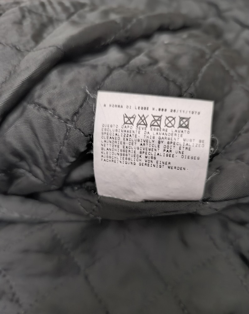 Puede incluir: Un interior de tela acolchada gris oscuro con una etiqueta de cuidado rectangular blanca. La etiqueta muestra el texto A NORMA DI LEGGE N.883 26/11/1973 seguido de cinco s&iacute;mbolos de lavado e instrucciones en italiano, ingl&eacute;s, franc&eacute;s y alem&aacute;n.
