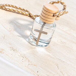 May include: A small, clear glass bottle filled with liquid, topped with a light-colored wooden cap. A silver-toned metal cross charm hangs from the neck of the bottle. A braided cord with gold-colored metallic threading is attached to the cap.