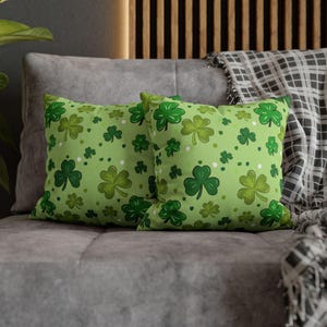 May include: Two square throw pillows with a light green background and a pattern of various sized shamrocks in shades of green. The pillows are resting on a gray sofa, with a plaid blanket visible in the background.