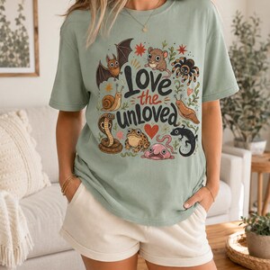 May include: A sage green short-sleeve t-shirt featuring a graphic print with the text "Love the unloved" surrounded by illustrations of a bat, mouse, spider, snail, slug, cobra, frog, worm, lizard, and blobfish, along with small red hearts and stars.