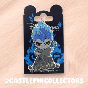 May include: A pin featuring Hades from Disney's Hercules, with blue flames and a dark gray outfit. The pin is on a black backing card with the Disneyland Paris logo and blue flame accents. The pin is a collectible item.