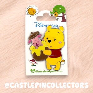 May include: A Disney pin featuring Winnie the Pooh and Piglet. Winnie the Pooh is yellow and wearing a red shirt. Piglet is pink and in a honey pot. The pin has a sun, tree, and bee design at the top. The Disneyland logo is visible.