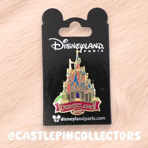 Può includere: Una spilla di Disneyland Paris con un design a castello colorato. La spilla è montata su una carta nera con il logo di Disneyland Paris e l'indirizzo del sito web. La spilla ha uno striscione rosso con la scritta "Disneyland Paris".