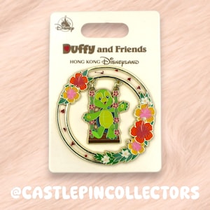 May include: A Duffy and Friends pin from Hong Kong Disneyland. The pin features a green turtle character on a swing within a floral wreath. The pin is on a white card with the Disney logo and text.