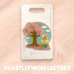 May include: Enamel pin featuring a cartoon character under a cherry blossom tree. The pin has a light blue background with white clouds. The character is wearing a blue outfit. The pin is on a white card with the text "Disney Parks Collection".