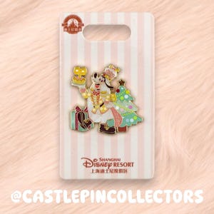 May include: A Shanghai Disney Resort enamel pin featuring Goofy dressed in a festive gold and white outfit, holding a small cake. The pin includes a decorated Christmas tree and a gift box, mounted on a white and pink striped backing card.