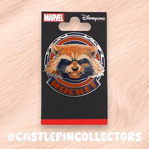 Op de afbeelding: Een ronde metalen emaille speld met het gezicht van het Marvel-personage Rocket Raccoon. De speld is geplaatst tegen een donkerblauwe en oranje cirkelvormige achtergrond met de naam ROCKET in het oranje gedrukt. Het zit op een zwarte kaart.