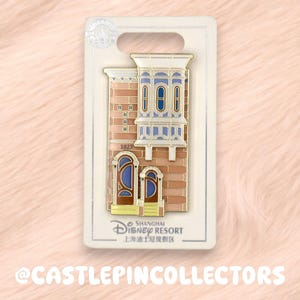 Könnte beinhalten: Ein Shanghai Disney Resort Emaille-Pin, der ein Gebäude aus beigefarbenen Ziegeln mit einem weißen Erkerfenster, blauen Bogentüren und der Zahl 1023 zeigt. Der Pin ist auf einer weißen Karte mit dem Disney-Logo und Text befestigt.