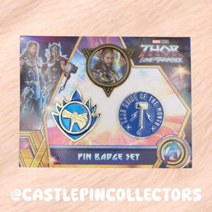 Könnte beinhalten: Ein Marvel Studios Thor Love and Thunder Anstecknadel-Set mit drei verschiedenen Pins auf einer Marken-Präsentationskarte. Das Set enthält einen runden Porträt-Pin, einen blau-goldenen schildförmigen Hammer-Pin und einen runden blauen Pin.