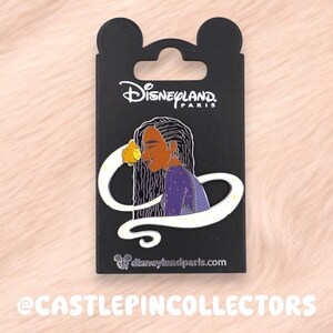May include: A Disneyland Paris enamel pin on a black backing card featuring a character with long dark hair wearing a purple top, holding a glowing yellow object. The card displays the Disneyland Paris logo and website address at the bottom.
