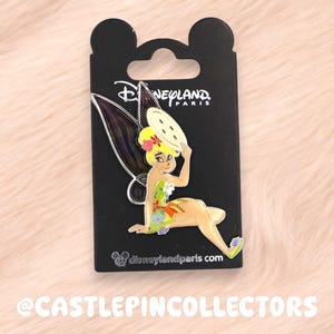 Op de afbeelding: Een Disney-pin met Tinkerbell, in een geel en groene jurk, die een bord vasthoudt. De pin zit op een zwarte kaart met het Disneyland Paris-logo en de website disneylandparis.com. De achtergrond is zacht, roze-beige.