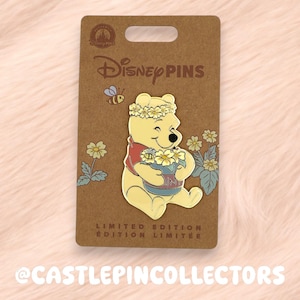 May include: A Disney pin featuring Winnie the Pooh holding a pot of flowers, wearing a flower crown, and smiling. The pin is on a brown card with the Disney Pins logo and the text "LIMITED EDITION". A small bee is flying above Pooh.