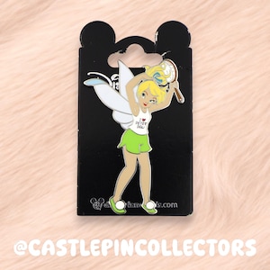 May include: A Tinkerbell enamel pin on a black backing card. The pin features Tinkerbell in a tennis outfit, holding a tennis racket and ball. She wears a white top with "I love Peter Pan" text and a green skirt. The pin has a Disney logo.