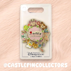 Peut inclure: Une broche en métal émaillé circulaire représentant les personnages de Duffy and Friends disposés autour d'un cercle rose central avec le texte Duffy and Friends. La broche est fixée sur une carte de support rectangulaire blanche.