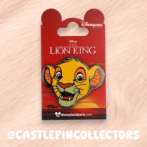 May include: A Disney The Lion King pin featuring Simba's face. The pin is on a red backing card with the movie title and Disneyland Paris branding. The background shows a sunset scene. The pin is yellow, orange, and green.
