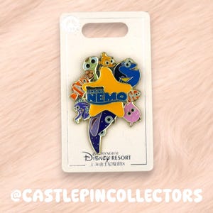 Op de afbeelding: Een kleurrijke emaille pin met personages uit de film "Finding Nemo". De pin bevat Nemo, Dory en andere personages, tegen een gele zeester. De pin is gemonteerd op een witte kaart met het logo van Shanghai Disney Resort.