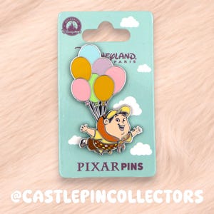 May include: A Pixar pin featuring a character from the movie "Up" floating with colorful balloons. The pin is on a light blue backing with the words "Disneyland Paris" and "Pixar Pins". The pin is set against a soft, peach-colored background.