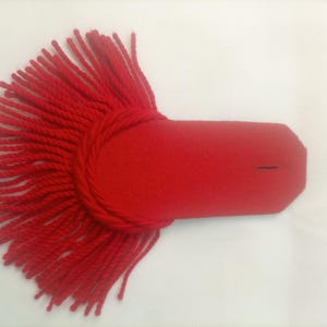 Puede incluir: Una charretera de color rojo brillante con una base de fieltro rectangular que termina en punta y tiene una pequeña ranura horizontal. El extremo redondeado está adornado con una densa franja de hilos de lana rojos en semicírculo.