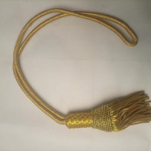 Könnte beinhalten: Ein goldfarbener Vorhanghalter mit einer langen, geflochtenen Kordelschlaufe, die an einem dekorativen, zylindrischen Quastenkopf befestigt ist, mit einer dichten Franse aus passenden goldfarbenen Fäden, die ein einheitliches metallisches Aussehen erzeugt.