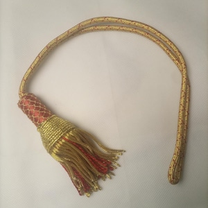 Puede incluir: Una borla decorativa con una parte superior cilíndrica roja cubierta por una malla dorada, unida a un cordón largo dorado. La base consiste en un denso grupo de hilos dorados y rojos colgantes, creando un efecto de flecos superpuestos.
