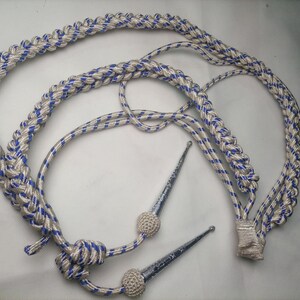 Puede incluir: Un cordón de aiguillette trenzado con un patrón repetitivo de hilos blancos y azules. El cordón incluye dos herretes de metal plateado con puntas afiladas y dos pequeños nudos decorativos blancos, redondos y texturizados.